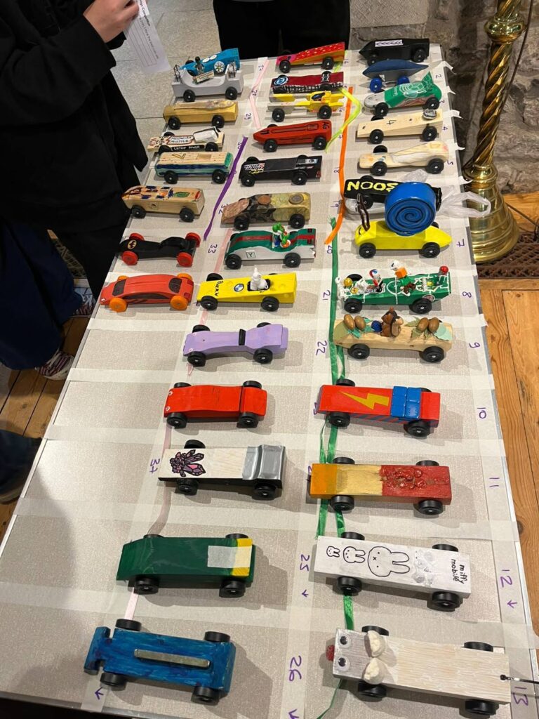 Rows of handmade painted pinewood derby cars lined up and numbered on a taped track, including snail, rocket and cartoon-themed designs.
